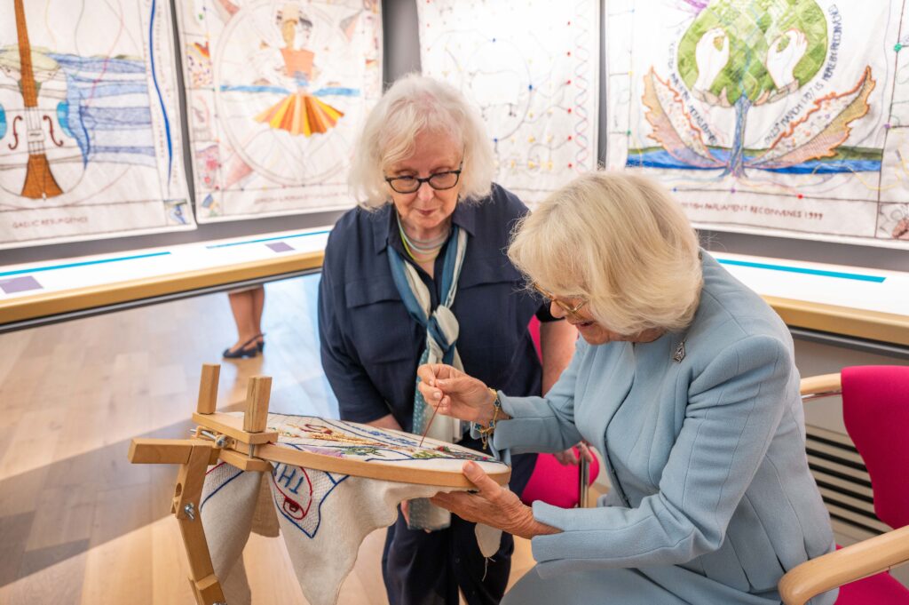 Dorie Wilkie, Head Stitcher, and Her Majesty, Queen Camilla put a stitch in a specially designed panel to mark the visit to the Great Tapestry by Their Majesties